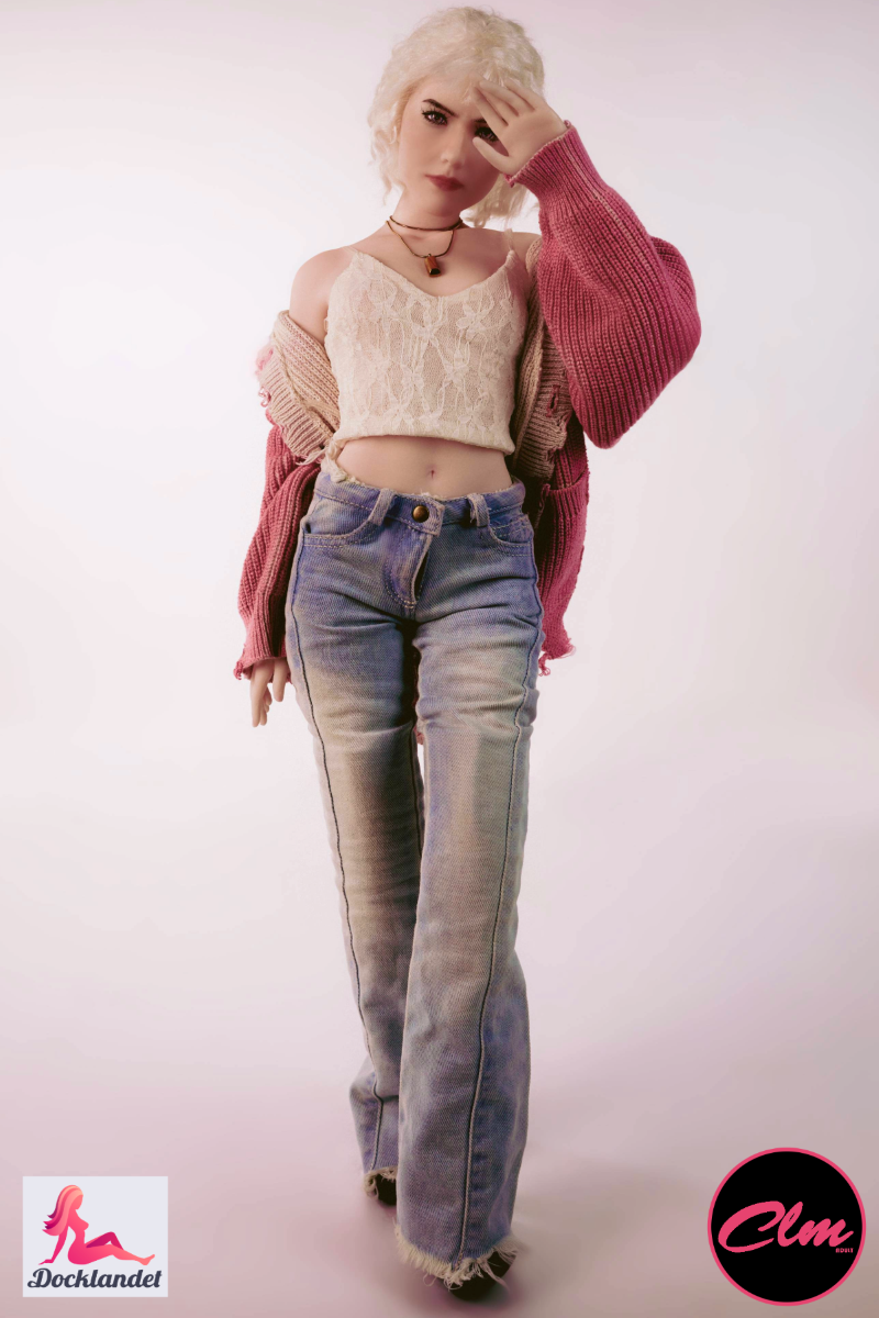 Boneca sexual Tara mini realista 60 cm em silicone, boneca loira de jeans, top curto e jaqueta rosa, em pé contra fundo claro de estúdio.